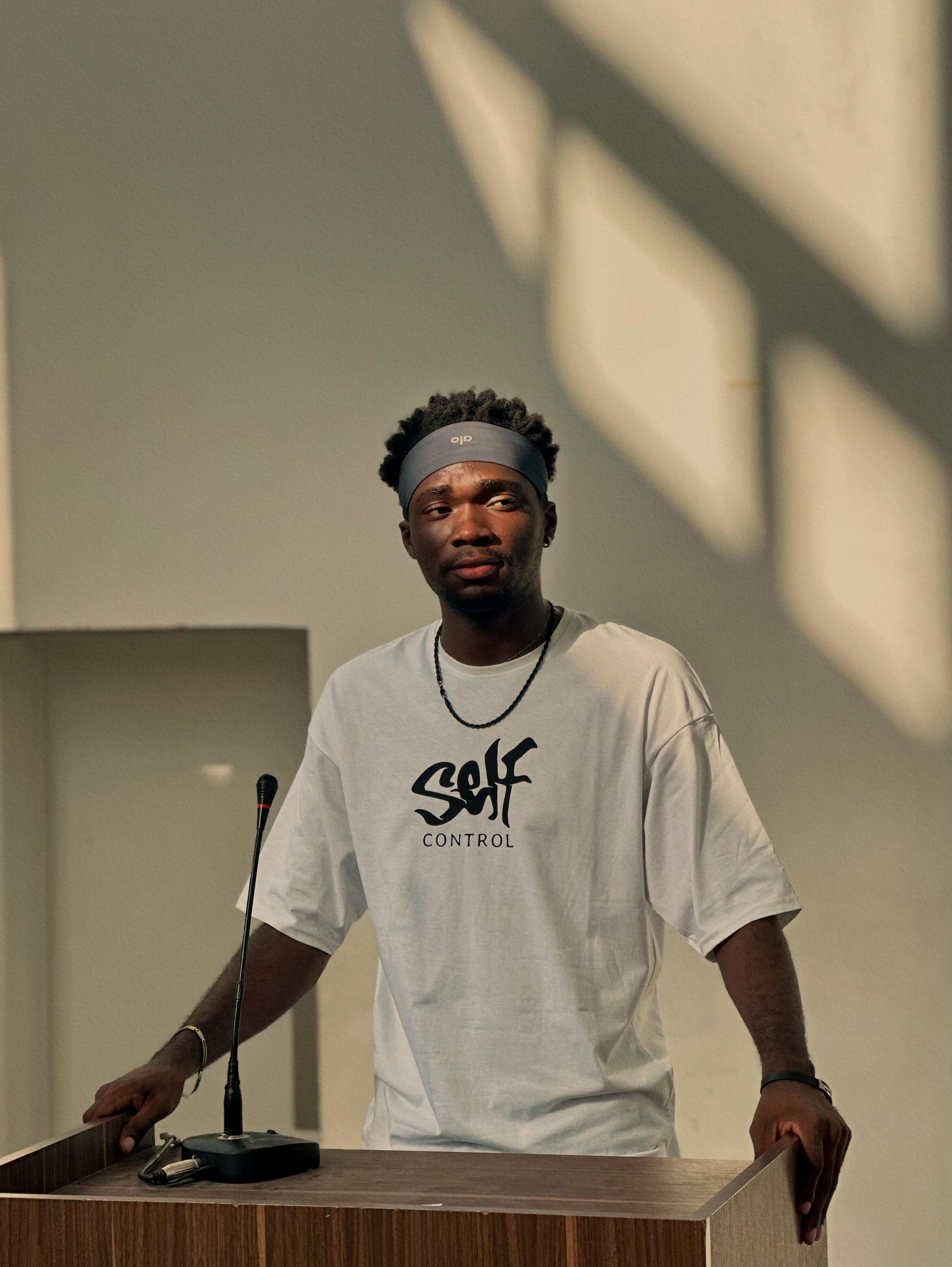 Young Black man in a white graphic t-shirt and headband stands behind a wooden podium.