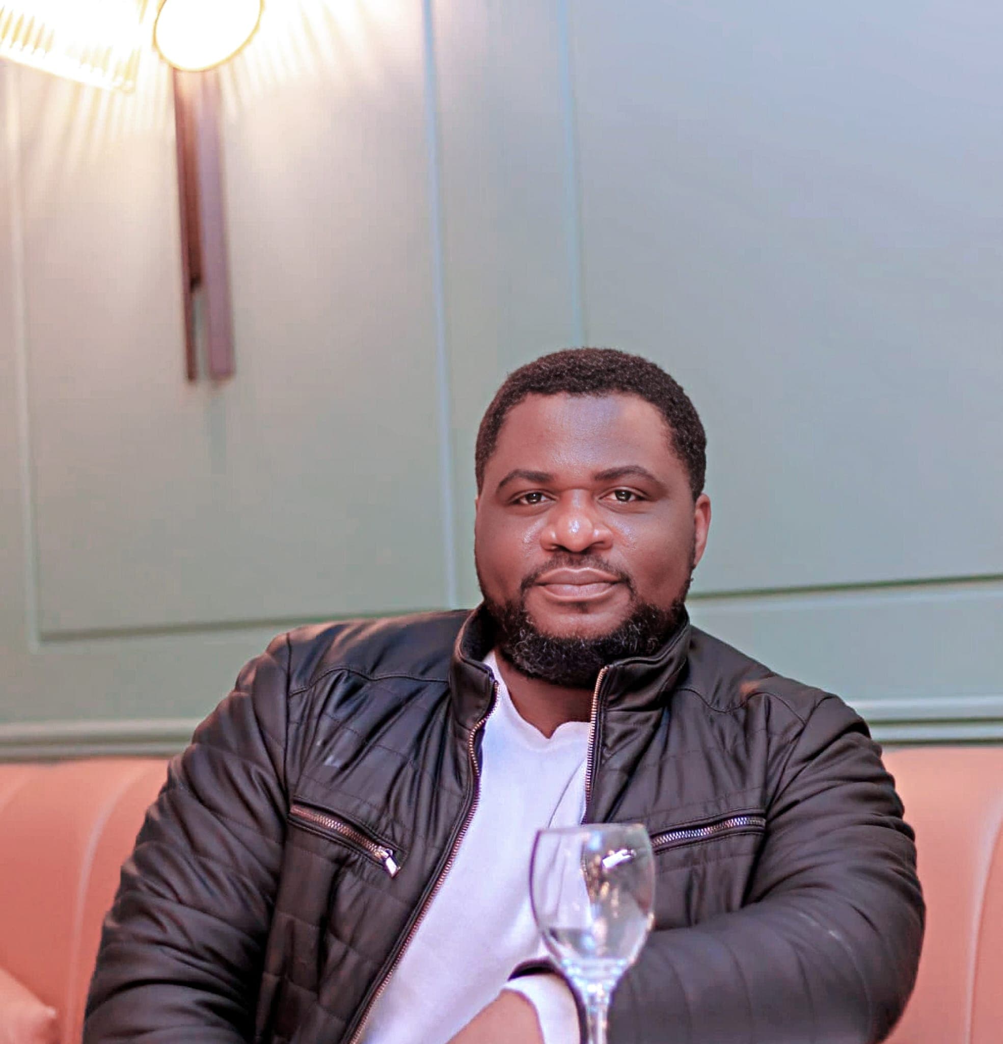 Bearded man wearing a black quilted jacket sits at a table with a wine glass.