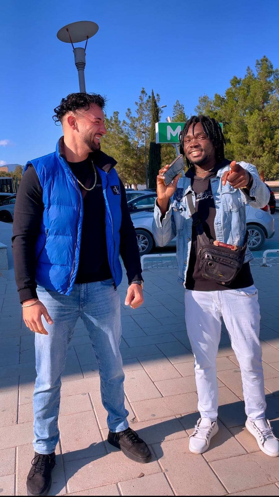 Two men smiling outdoors, one in a blue vest and another pointing at the camera.