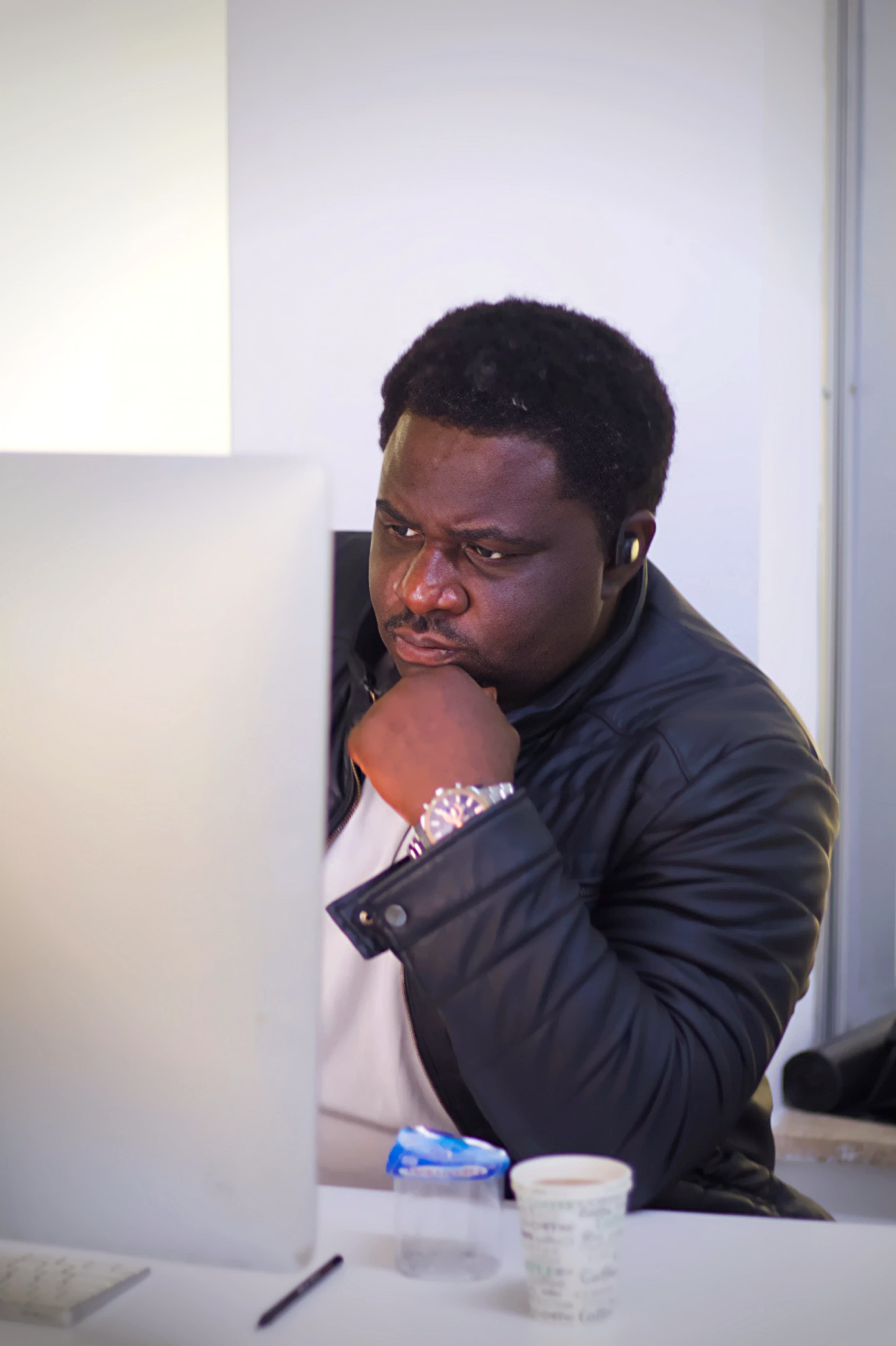 Focused Black man in a leather jacket stares intently at a computer screen, hand on chin.
